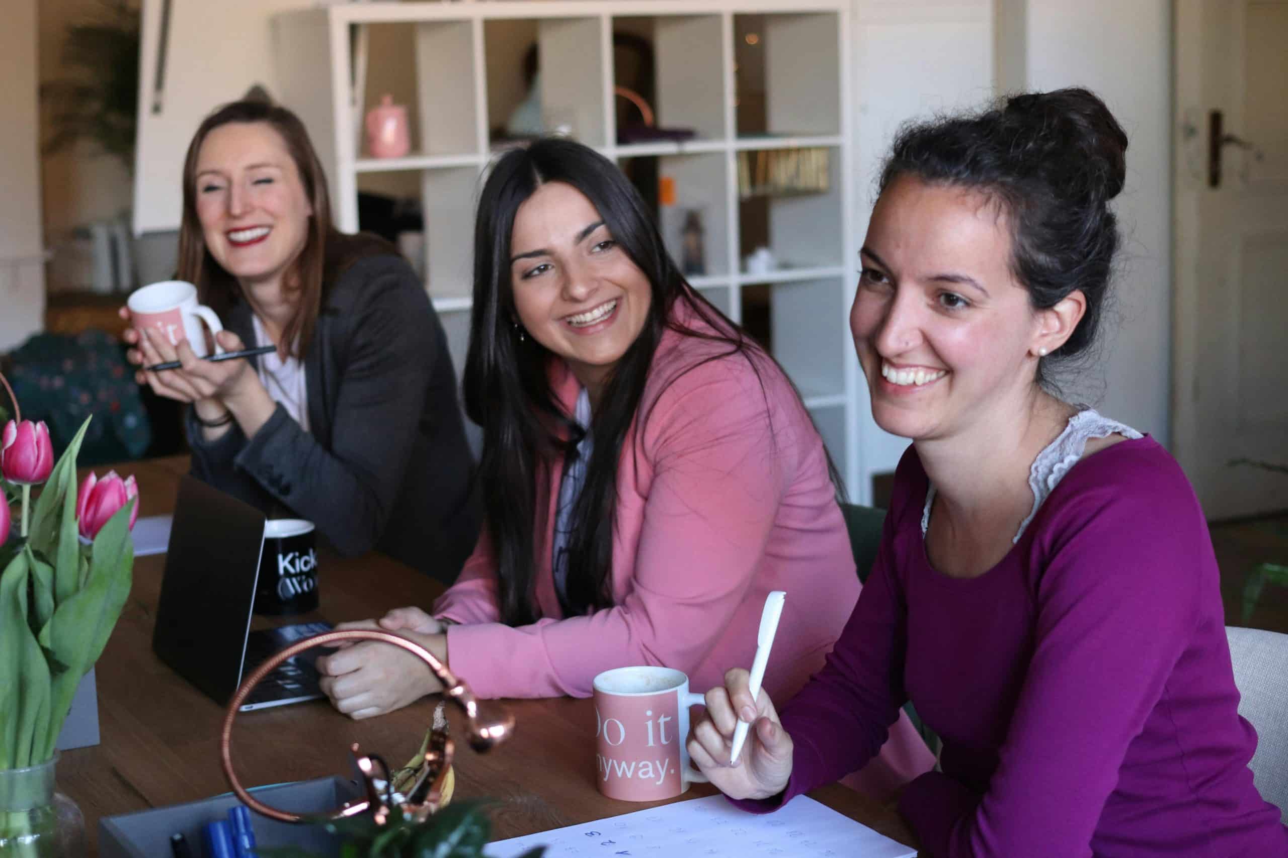 Drie vrouwen zitten lachend aan tafel en drinken koffie